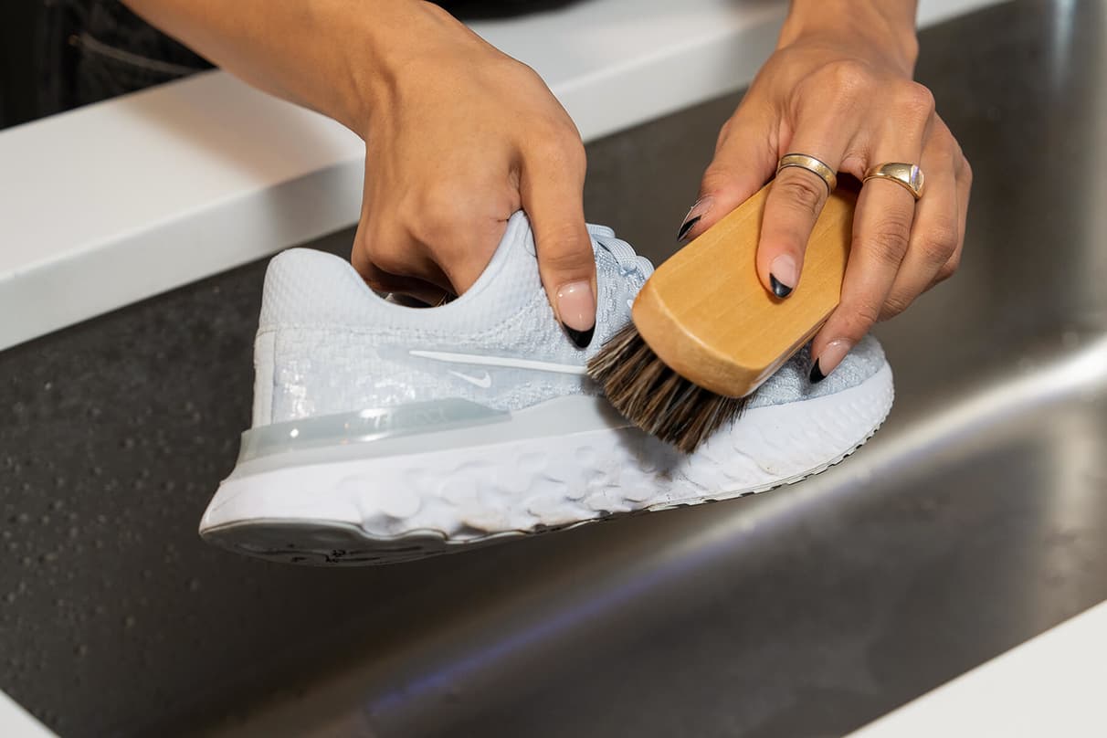 Hands holding a white mesh Nike shoe while a soft brush cleans the knit upper