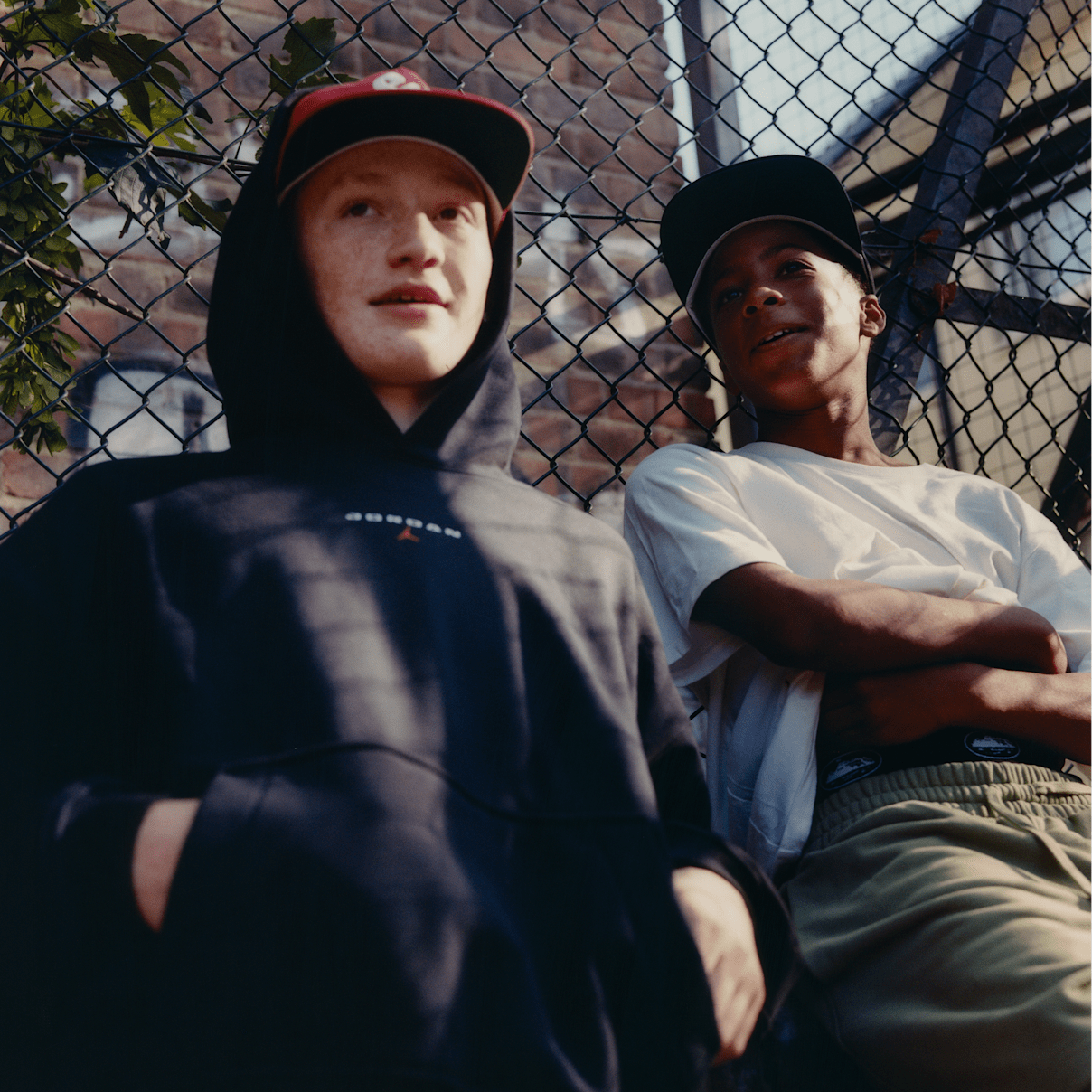 Two boys sitting against a chain-link fence, one wearing a black Jordan hoodie and cap, the other in a white t-shirt and cap., MECCSNAPI ALAPDARABOK