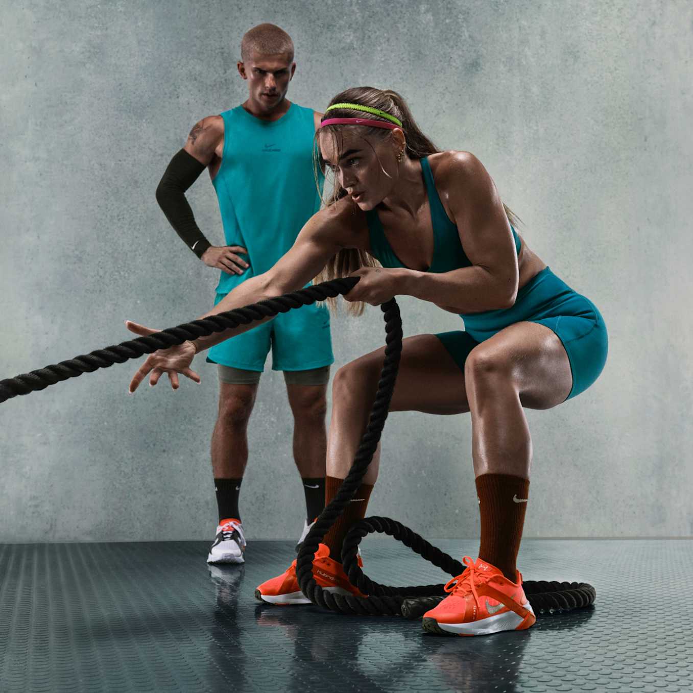 En kvinne som står i knebøy og trekker i et kamptau i et treningsstudio. Hun har på seg en blå Nike-sports-BH, shorts og oransje Nike-treningssko., Matchende sett