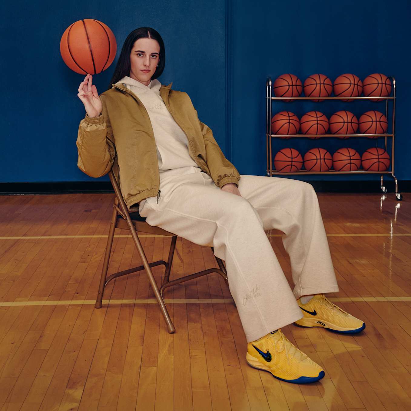 Caitlin Clark, bej bir eşofman ve sarı Nike Caitlin 1 ayakkabılarla bir sahada sandalyede oturmuş, basketbol topunu döndürüyor. , Caitlin 1 Özel Koleksiyonu