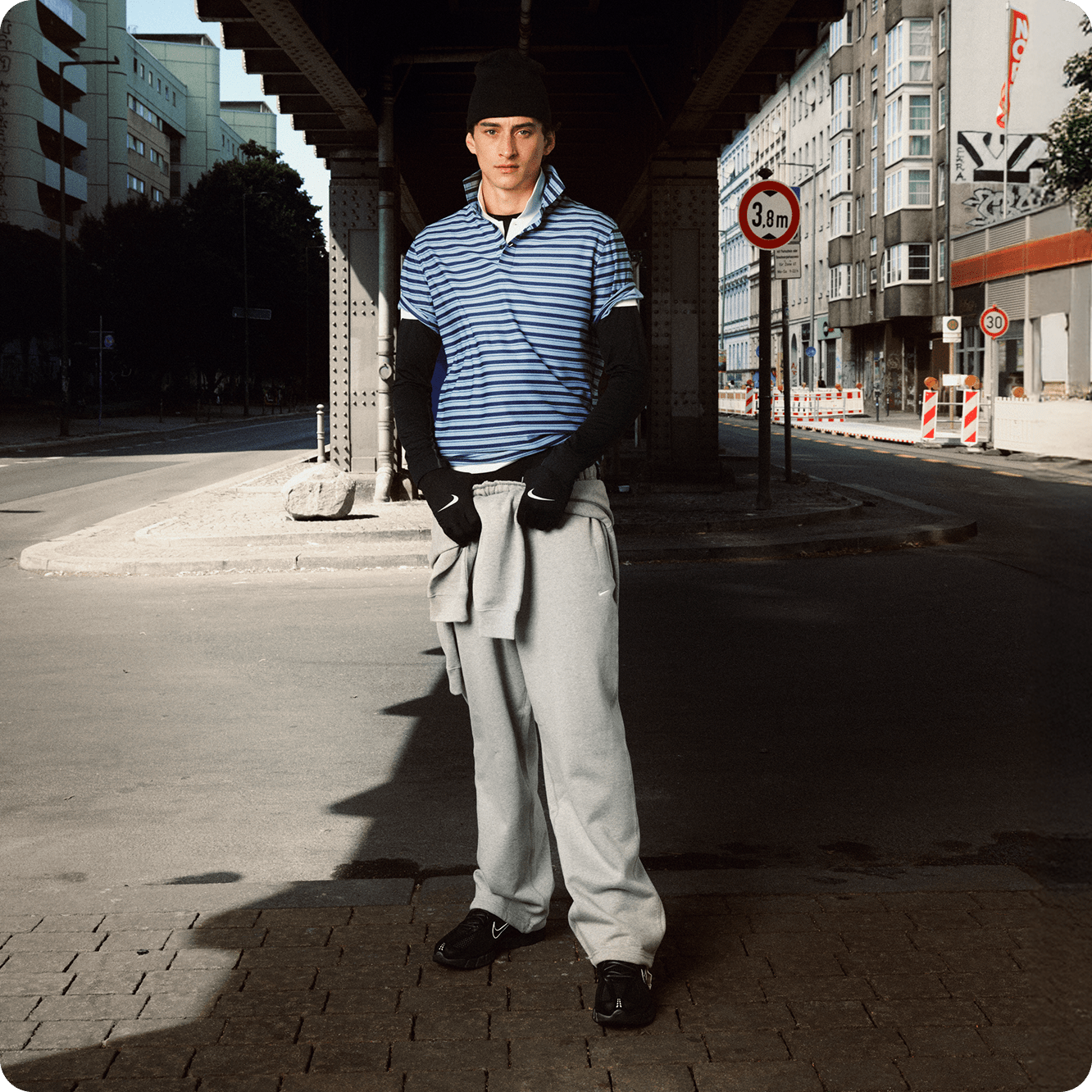 Ein Mann steht unter einer Brücke auf einer Straße. Er trägt eine schwarze Beanie, ein blau gestreiftes Poloshirt, ein schwarzes Langarmshirt, eine graue Nike Jogginghose und schwarze Nike Zoom Skylon 11 Sneaker., London