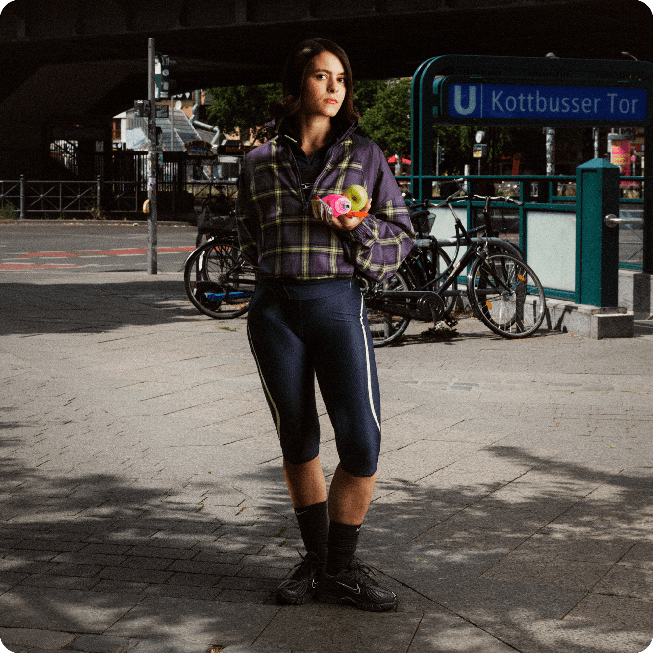 Een vrouw staat op een straat in de stad, met een appel en een roze waterfles in haar hand. Ze draagt een paars geruite jas, donkerblauwe cropped leggings en zwarte Nike Zoom Skylon 11-schoenen., Berlijn