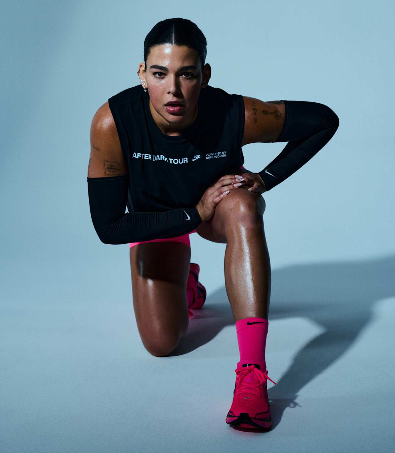 A woman kneeling with hands on her knee in a grey studio space, wearing a black sleeveless top, black arm sleeves, pink shorts, pink socks, and pink running shoes.