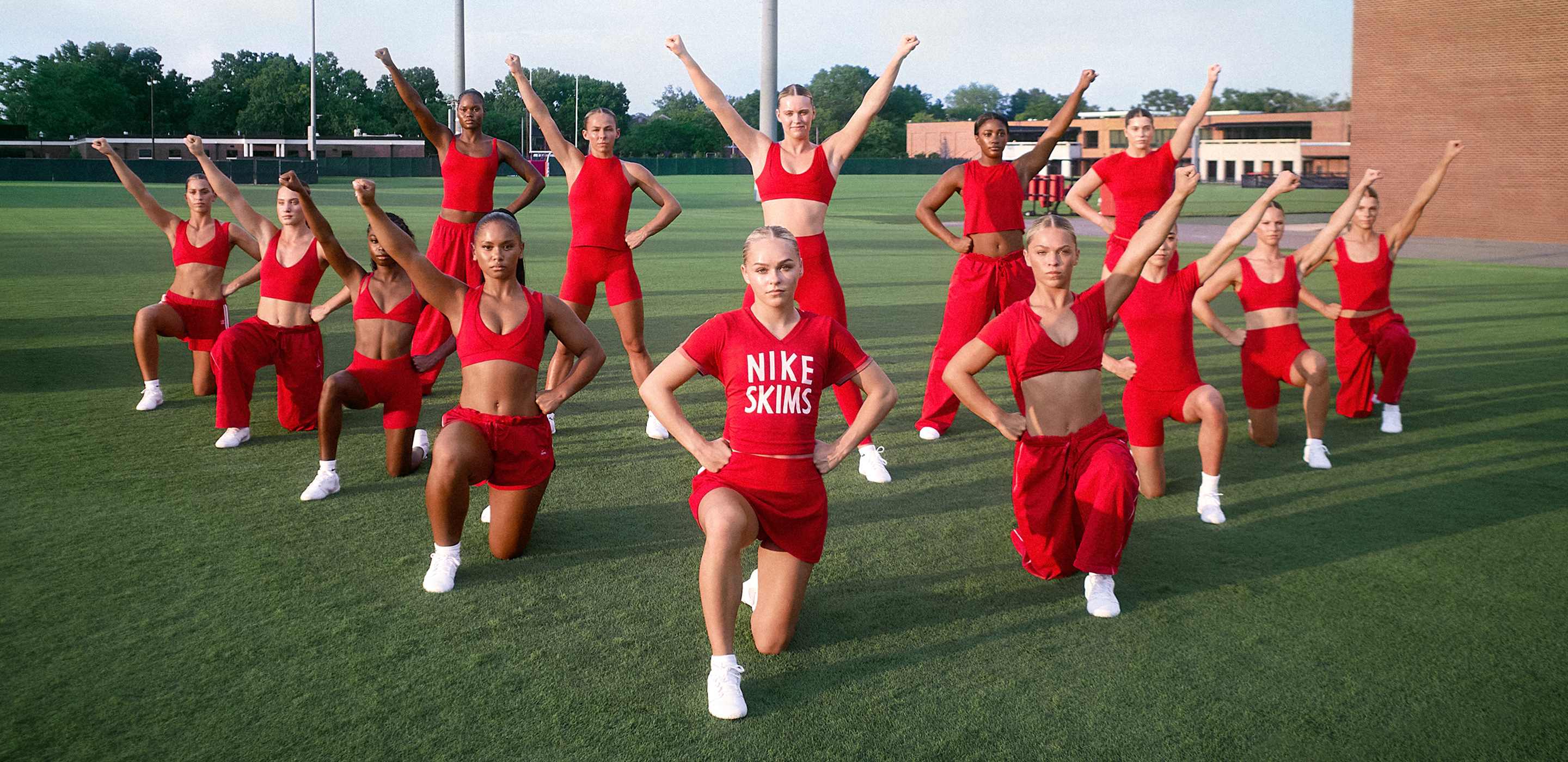 Een cheerleaders-team in een formatie van twee rijen, allemaal gekleed in rode NikeSKIMS. De voorste rij knielt, de achterste rij staat met een of twee armen omhoog.
