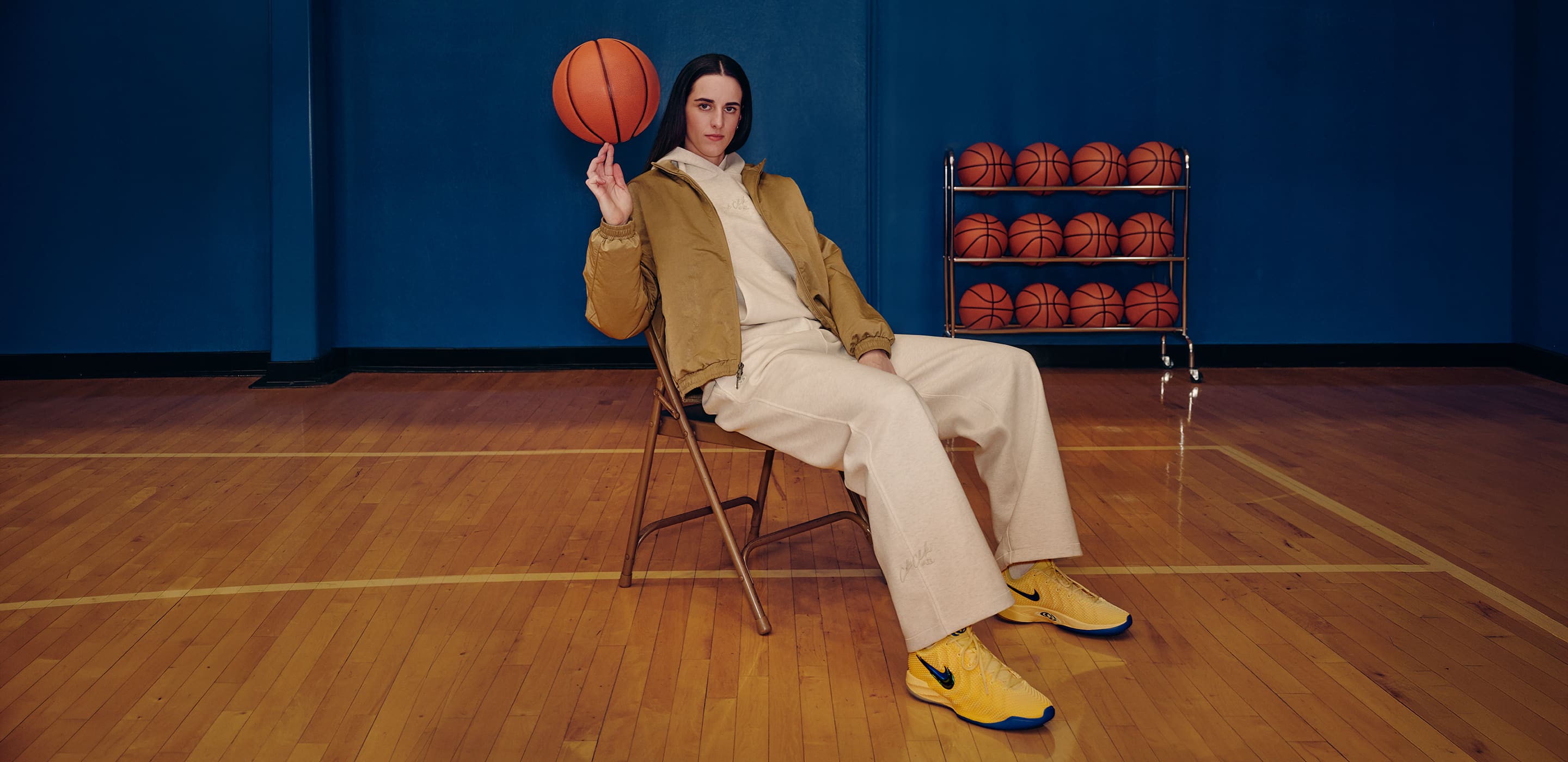Caitlin Clark egy széken ülve pörgeti a kosárlabdát a pályán. Bézs színű tréningruhát és sárga Nike Caitlin 1 cipőt visel. , Caitlin 1 egyedi kollekció