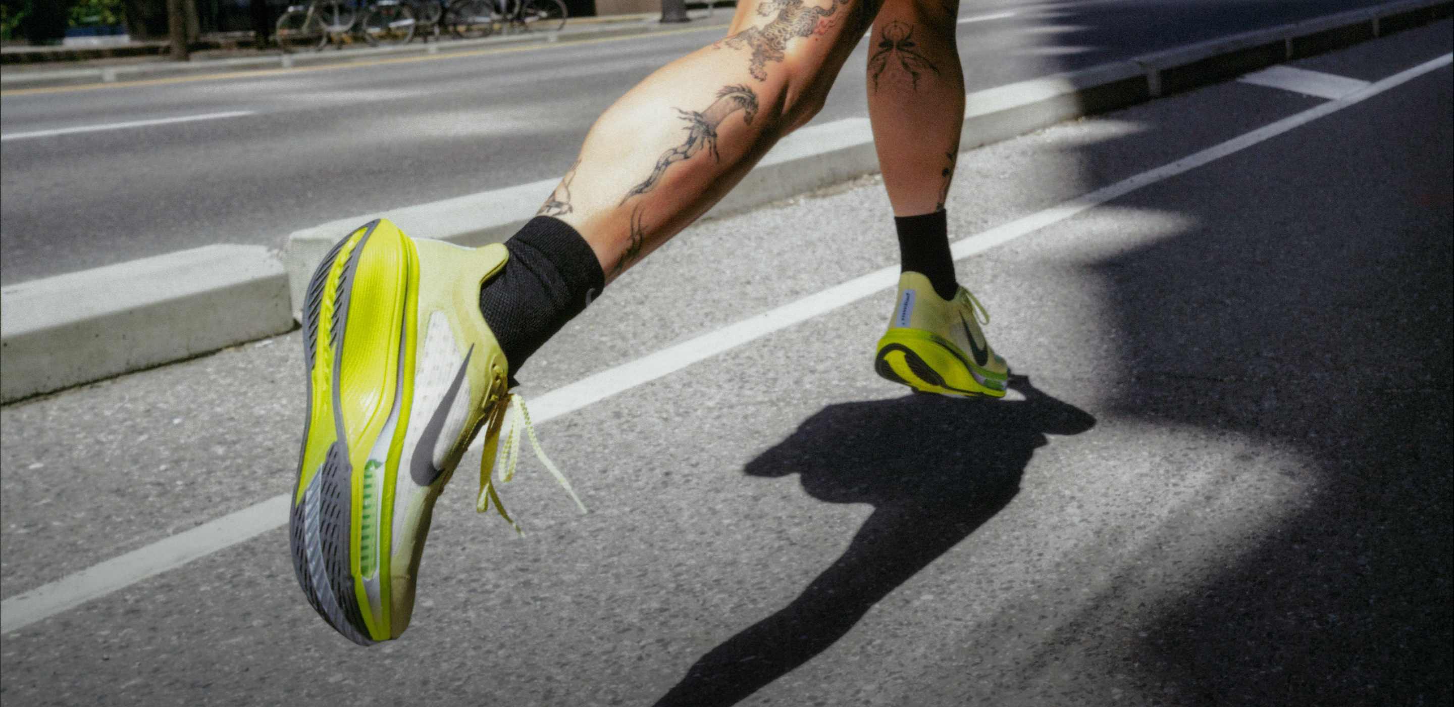 Una mujer corriendo por una calle urbana, con pantalones cortos negros, una camiseta negra y zapatillas de running Nike de color amarillo neón.