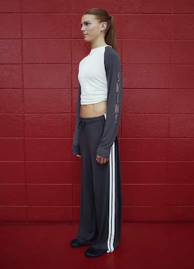 An athlete stood facing the right, wearing gray and white NikeSKIMS long-sleeved top and trousers, stood against a red wall.