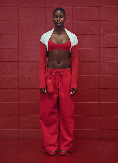 An athlete stood facing forward wearing a red NikeSKIMS bra, long-sleeved top, and pants against a red wall.