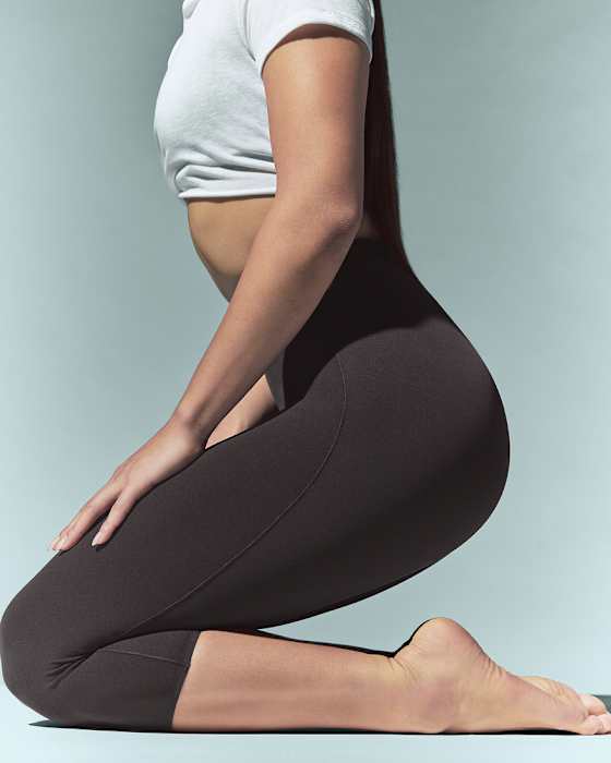 An athlete from the shoulders down, kneeling on the floor, wearing brown NikeSKIMS leggings, and a cropped white t-shirt., slide 2 of 7