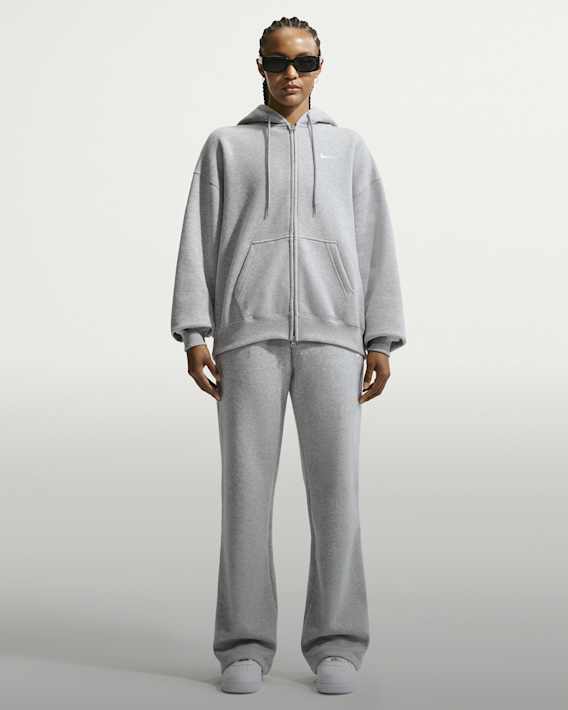 What’s New Now, A woman standing against a plain background, wearing a grey Nike zip-up hoodie, matching flare trousers, white trainers, and black sunglasses., slide 3 of 4