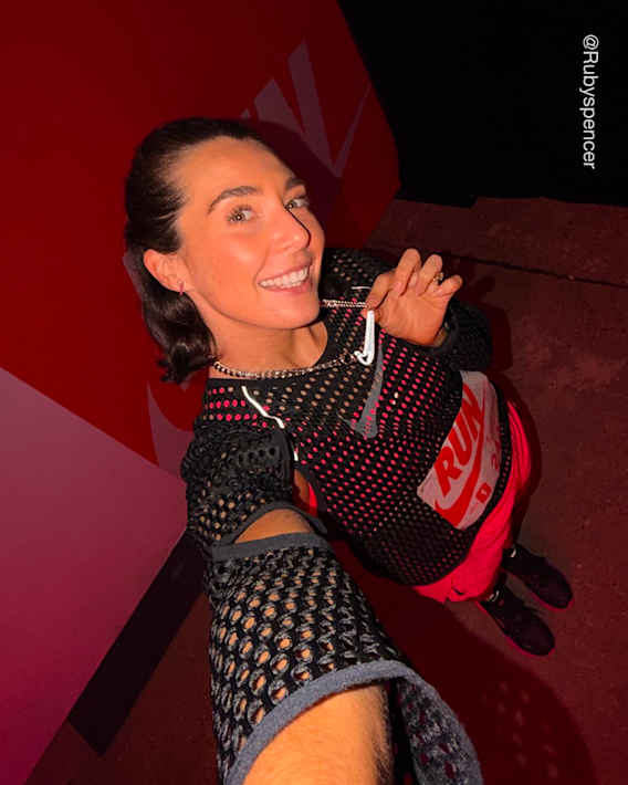 Uma mulher a sorrir e a levantar dois dedos num espaço interior com uma parede vermelha da Nike, vestindo uma camisola preta de rede com um dorsal "RUN" e calções cor-de-rosa., slide 1 of 3
