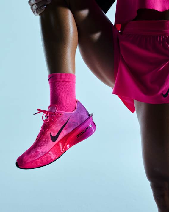 A person stretching in a studio space, wearing a pink Nike running shoe with a pink and purple gradient and pink athletic apparel., slide 2 of 3