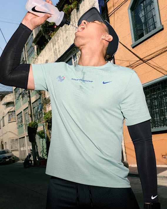 Győzd le a meleget, Egy férfi átlátszó Nike vizespalackból iszik az utcán, aki mentazöld Nike futópólót fekete rövidnadrágot, fekete karvédőt és fekete sapkát visel., slide 1 of 5