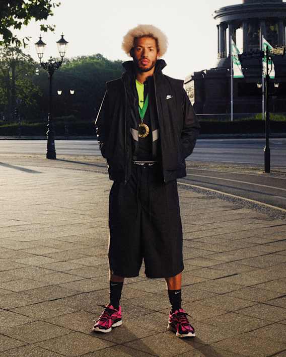 Man standing in an outdoor urban plaza wearing a black Nike outfit with pink Skylon Zoom 11 shoes and a large medal around the neck., slide 2 of 3