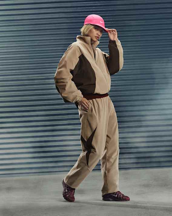 Full-body portrait of a women wearing a beige and brown Nike tracksuit, pink cap and dark red P-6000 sneakers, against a metallic urban backdrop., slide 3 of 3