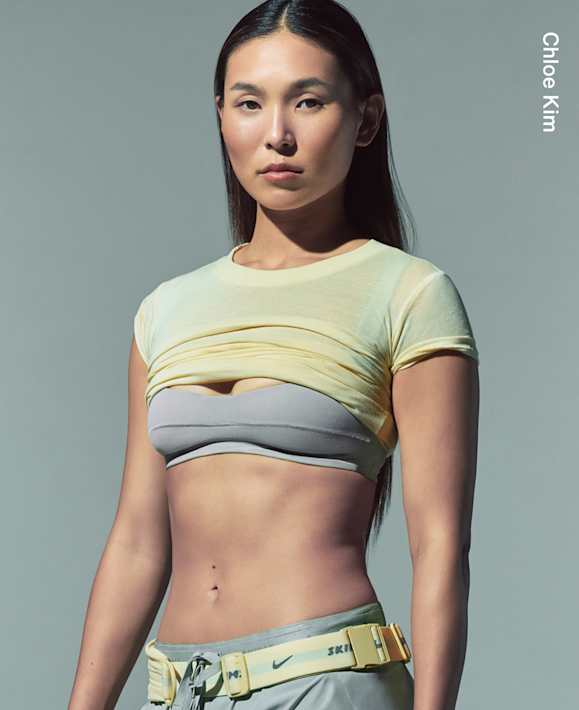 Getragen von Athletinnen, Chloe Kim steht in einem hellgrauen Studio und trägt ein kurzes gelbes T-Shirt, einen grauen Sport-BH und eine graue Hose mit gelbem NikeSKIMS Bund., slide 3 of 3