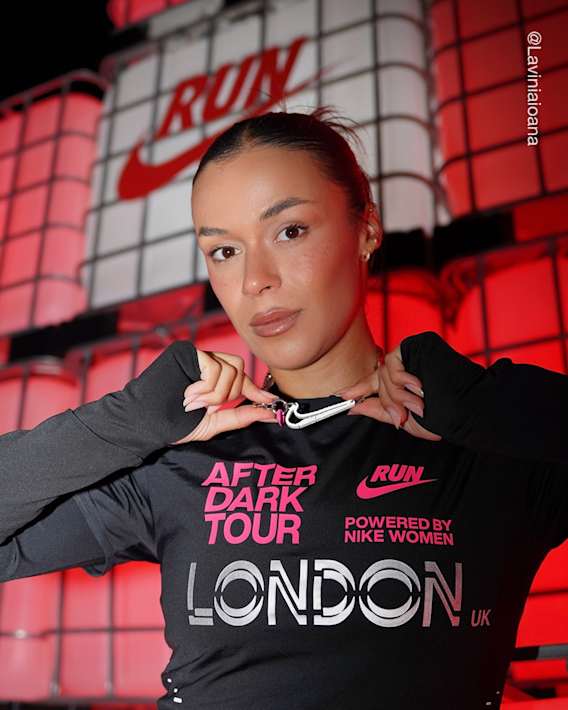Een vrouw staat tegen een rood verlichte achtergrond met het Nike Run-logo, houdt een witte Nike Swoosh-hanger vast en draagt een zwarte top met lange mouwen en de tekst 'After Dark Tour London UK'., slide 3 of 3