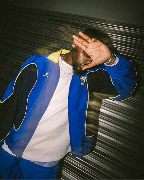 Trending Now, A man covers his face with his hand in front of a metal door wearing a jacket top of the blue and yellow Brasil Futebol x Jordan Brand collection., slide 1 of 4