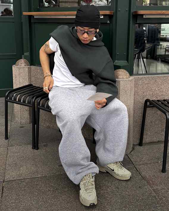 Trending, Een man staat voor een groene muur. Hij draagt een zwarte Nike pet, een donkergroen T-shirt, zwarte shorts, een lichtgrijze rugzak en donkergroene Nike mules., slide 2 of 5