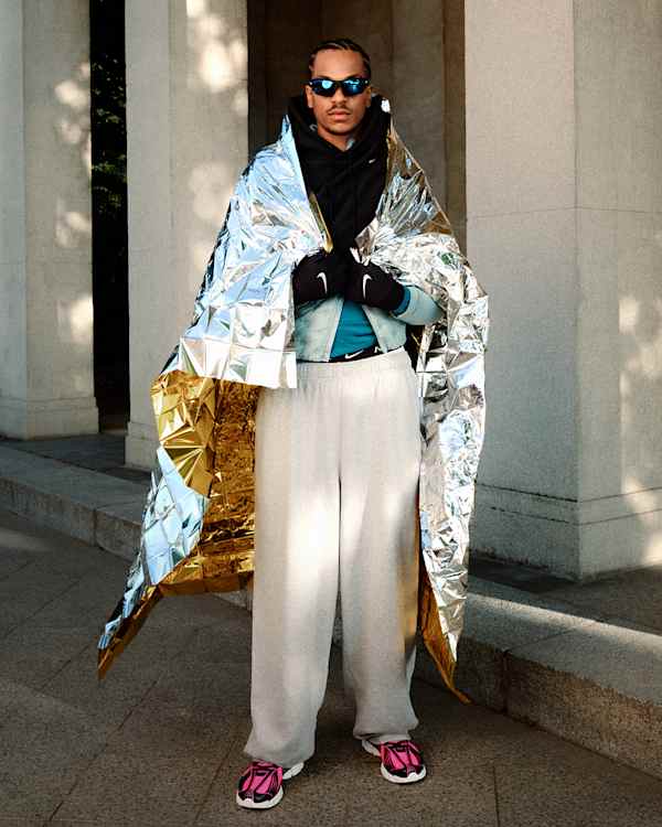 I trend del momento, Uomo in piedi davanti a colonne bianche, avvolto in una coperta termica oro e argento metallizzato, con pantaloni grigi e scarpe Nike Zoom Skylon 11 rosa e nere., slide 5 of 5