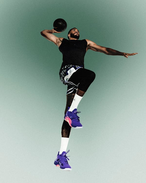 Trending, Een atleet hangt in de lucht terwijl hij zich klaarmaakt om een zwarte basketbal te gooien. Hij draagt een zwarte sportoutfit, witte sokken en paarse sneakers tegen een zachtgroene achtergrond., slide 3 of 4
