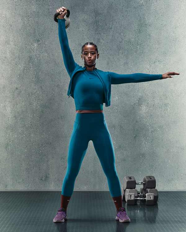 Shop by Activity, A woman lifting a kettlebell overhead in a gym with dumbbells on the floor, wearing a teal long-sleeve top, matching leggings, and purple trainers., slide 3 of 4