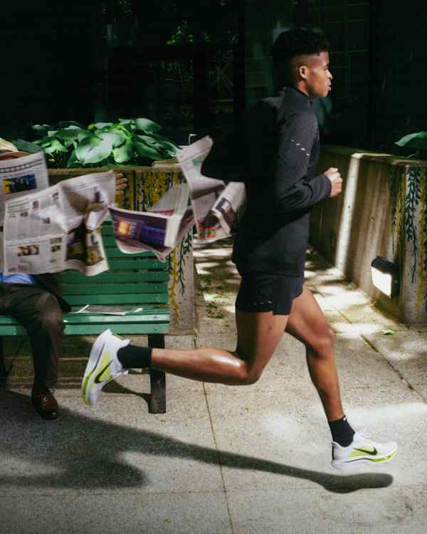 Most népszerű, Egy férfi fut egy árnyékos szabadtéri úton, fekete futóruházatot és fehér és neon sárga Pegasus Plus 2 cipőt visel., slide 1 of 4