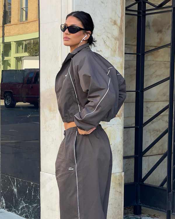 NikeSKIMS Styled By You, Model La’Tecia poses in a city setting wearing a NikeSKIMS tracksuit in Boulder and black sunglasses., slide 4 of 5