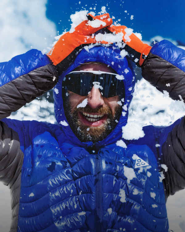 Trending Now, A man smiling with snow on his face and hands raised to his head in a snowy outdoor environment, wearing a blue ACG puffer jacket, orange gloves, and sunglasses., slide 2 of 5