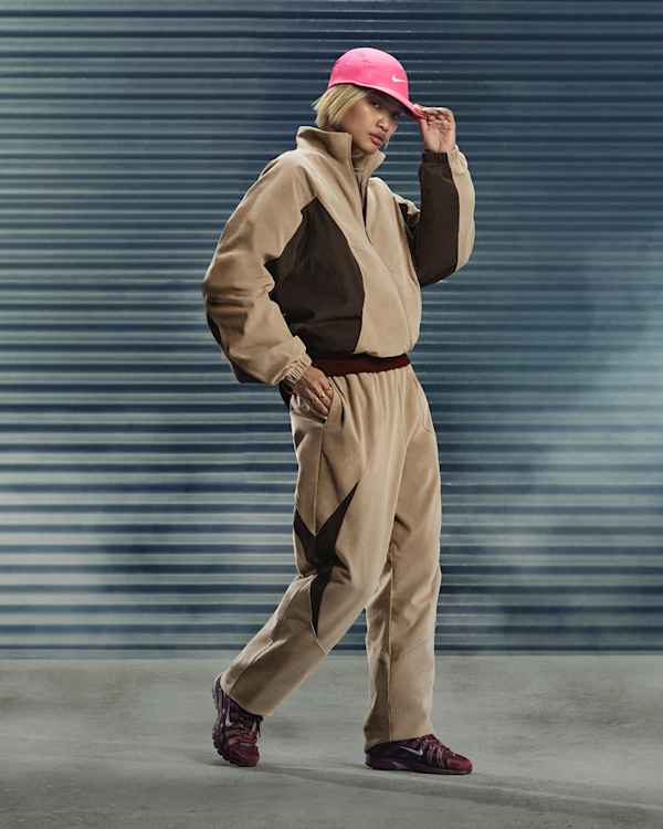 Wat is er nieuw, Een vrouw staat voor een grijs rolluik met haar hand op een roze Nike-pet. Ze draagt een beige fleece trainingspak en donkerrode Nike P-6000-sneakers., slide 1 of 5