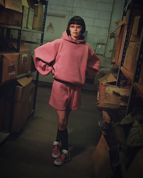 Wat is er nieuw, A woman standing in a warehouse, wearing a pink Jordan hoodie, matching shorts, and red and white Jordan shoes., slide 3 of 4