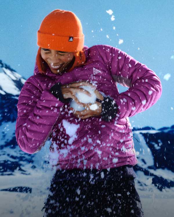 Co nowego?, Uśmiechnięta osoba w pomarańczowej czapce i fioletowej kurtce typu puffer Nike ACG Lava Flow trzyma śnieżkę, a wokół niej rozpryskuje się śnieg., slide 4 of 5
