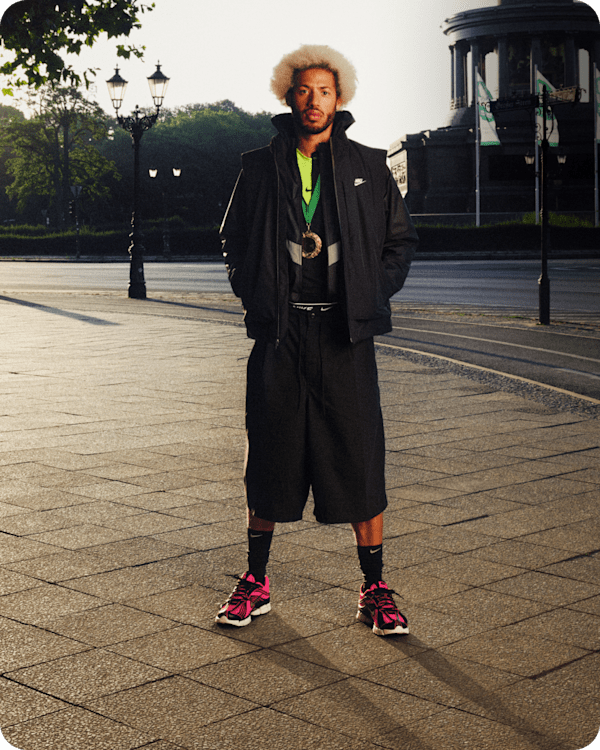 Un homme portant une veste Nike noire, un short ample, une médaille d'or et des baskets Nike Zoom Skylon 11 rose vif et noires, debout à l'extérieur., slide 3 of 3