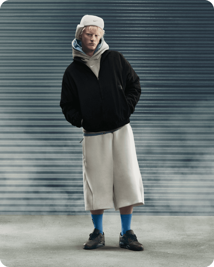 Mehr Inspo für deine Nike Sportswear Looks, Ein Mann steht vor einem grauen Rollladen und trägt eine helle Kappe, einen grauen Hoodie, eine schwarze Jacke, hellgraue Shorts, blaue Socken und dunkle Nike Air Max Sneaker., slide 4 of 5
