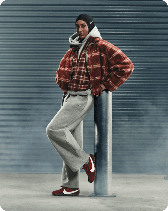 Mehr Inspo für deine Nike Sportswear Looks, Eine Frau lehnt an einer Metallsäule vor einer grauen Wand. Sie trägt eine schwarze Sturmhaube, eine rot karierte Jacke, einen grauen Trainingsanzug und rot-weiße Nike Moon Shoes., slide 2 of 5
