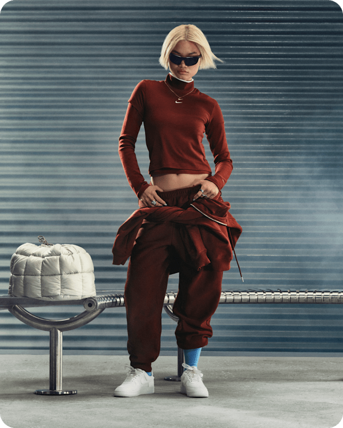 Meer manieren om Nike Sportswear te stylen., Een vrouw staat bij een metalen bank tegen een grijze golfplaatwand. Ze draagt een donkerrode coltrui, een bijpassende broek, een zwarte zonnebril en witte Nike Air Force 1-sneakers., slide 3 of 5