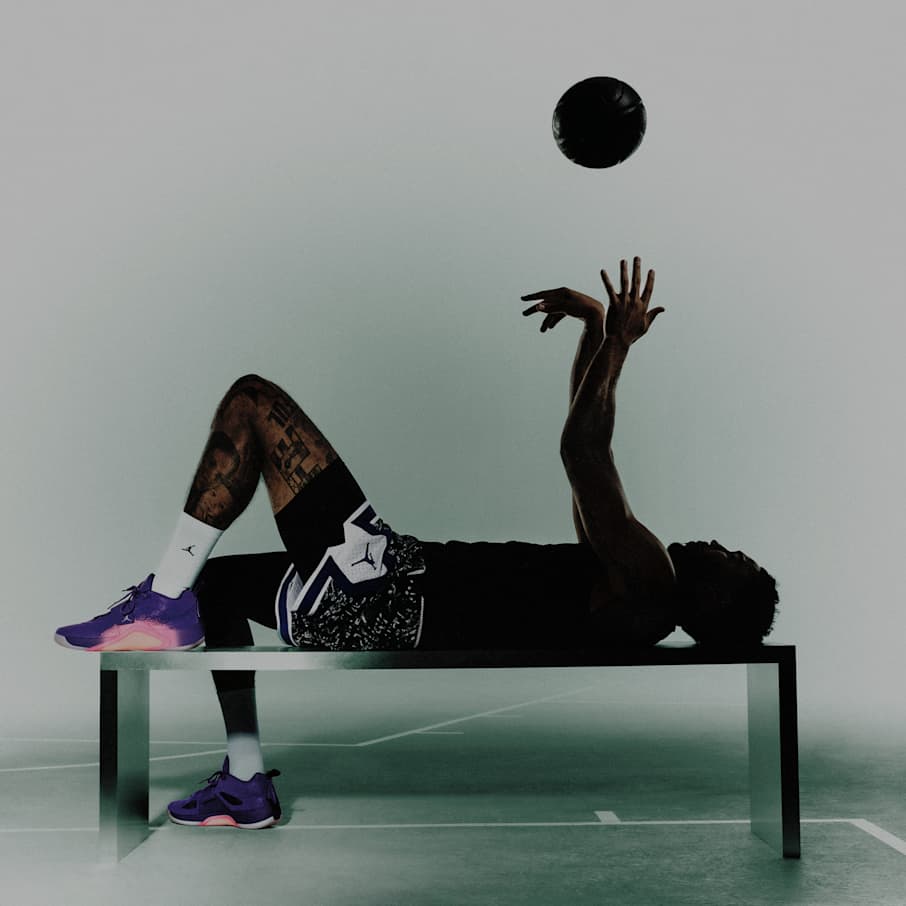 Ein Athlet liegt auf einer Bank und wirft einen schwarzen Basketball über den Kopf. Er trägt ein schwarzes Sportoutfit, weiße Socken und lila Sneakers und befindet sich in einem Trainingsraum in einer Halle., SICHERER HALT. EXPLOSIVE LIFT-OFFS.