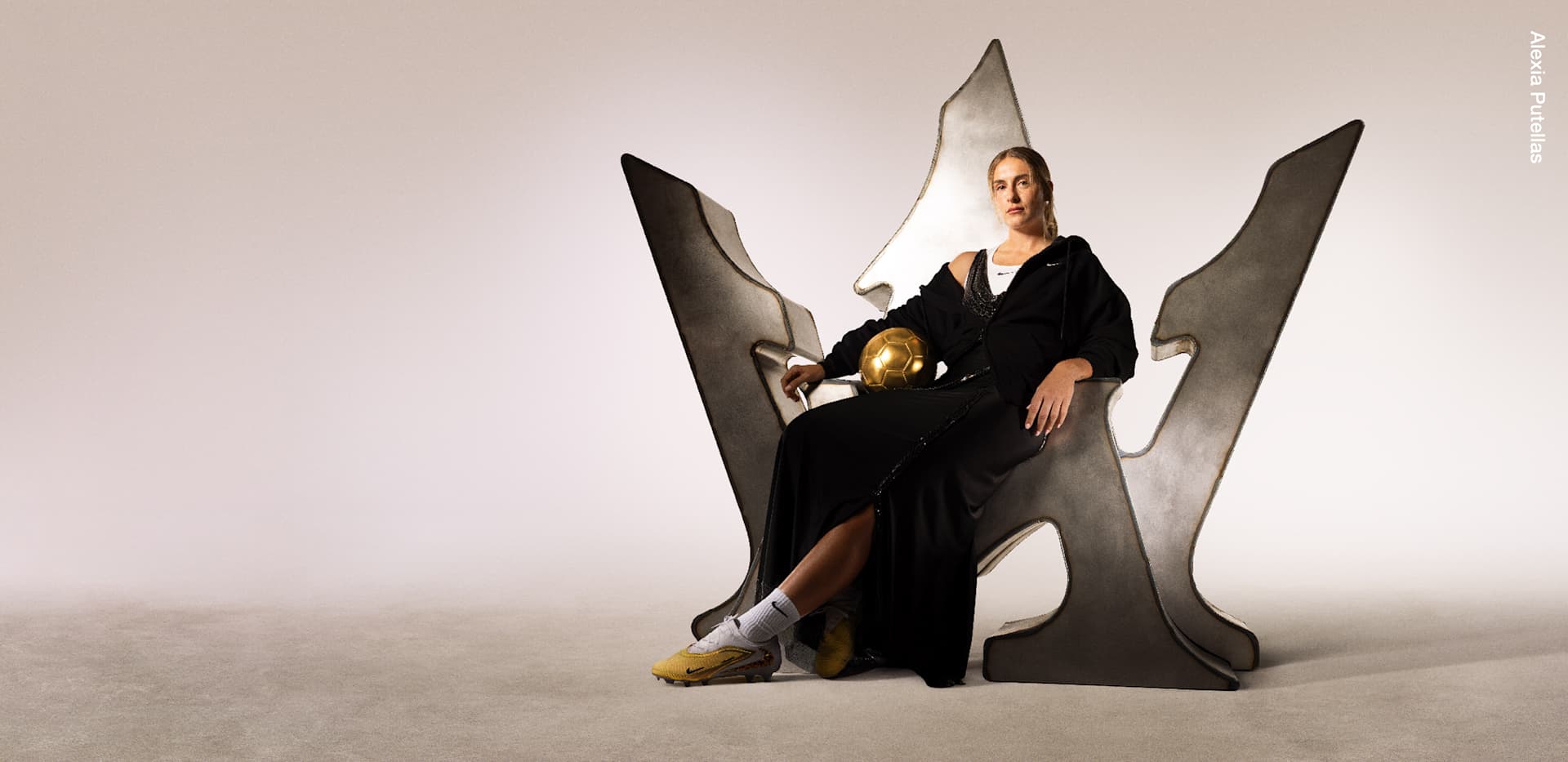 Alexia Putellas sits on a metallic throne shaped like her personal monogram, combining the letter “A” with two number 1s to form a crown-like silhouette. She wears Nike apparel and gold football boots, with one foot resting on a gold football in a portrait celebrating her Ballon d'Or achievement., THE THRONE IS HERS.
