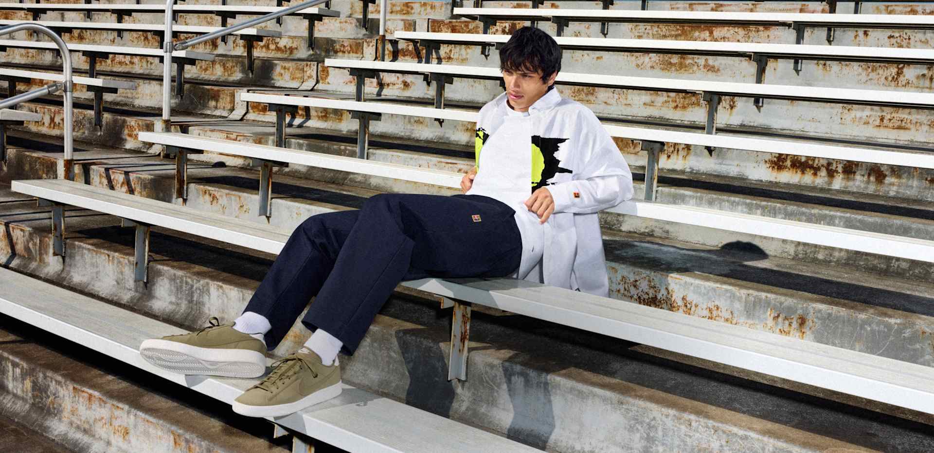 Man reclining on stadium bleachers wearing a white graphic shirt, navy pants, white socks, and olive green Nike Tennis Classic sneakers., READY FOR <br>ANYTHING