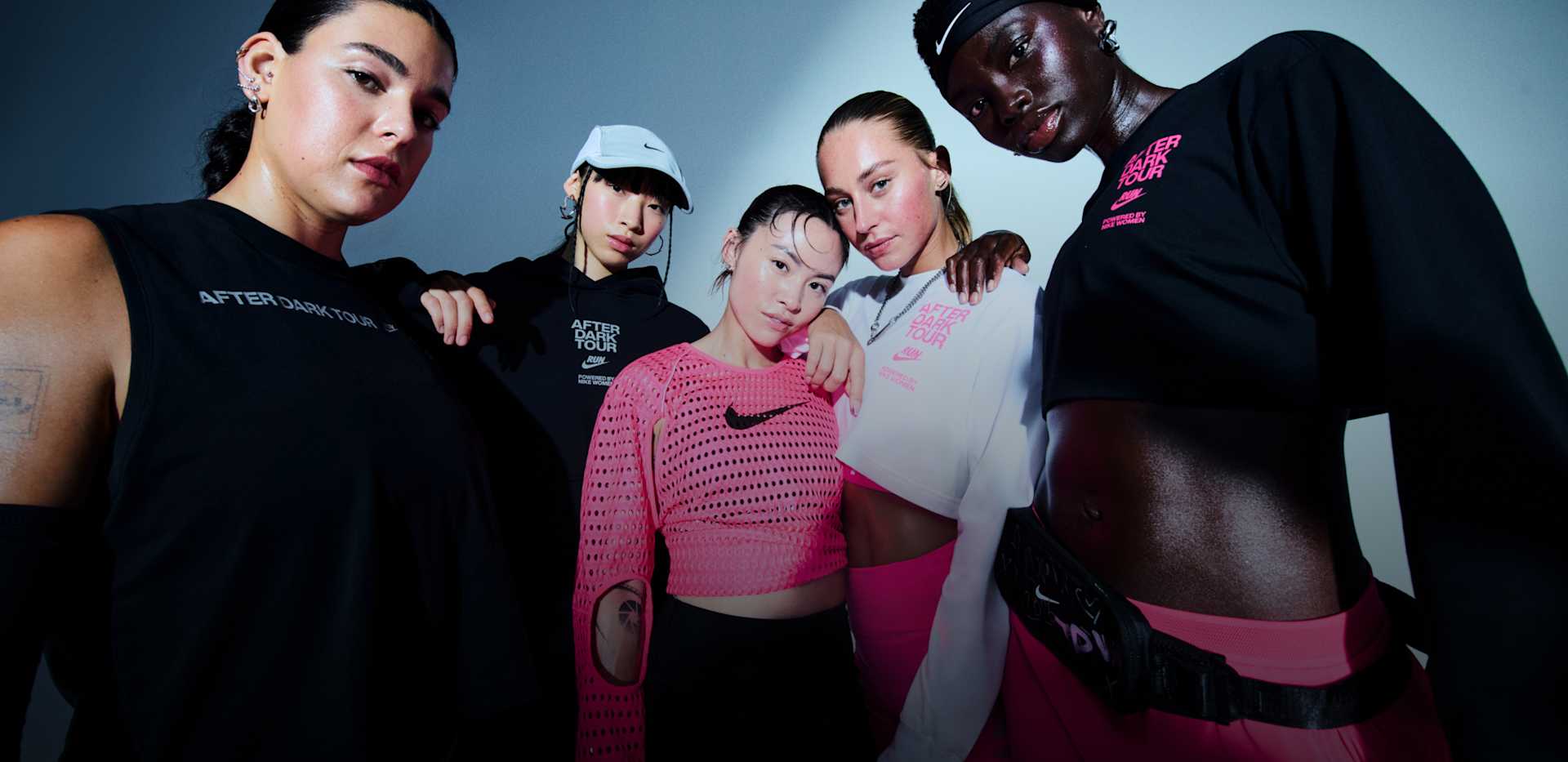 Five women standing and posing in a studio space, wearing Nike After Dark Tour Collection apparel, with one wearing a light blue hat and another a black waist bag., THIS RACE IS<br>PERSONAL