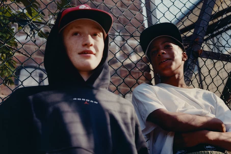 Deux jeunes hommes assis contre un grillage, l'un portant un sweat à capuche et une casquette Jordan noirs, l'autre un t-shirt blanc et un short kaki., À PORTER SANS MODÉRATION