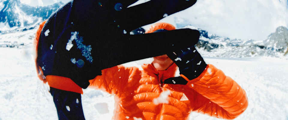 En person i en knalloransje Nike ACG Lava Flow-boblejakke har på hansker og strekker hendene mot kameraet, med et snødekt fjellandskap i bakgrunnen.