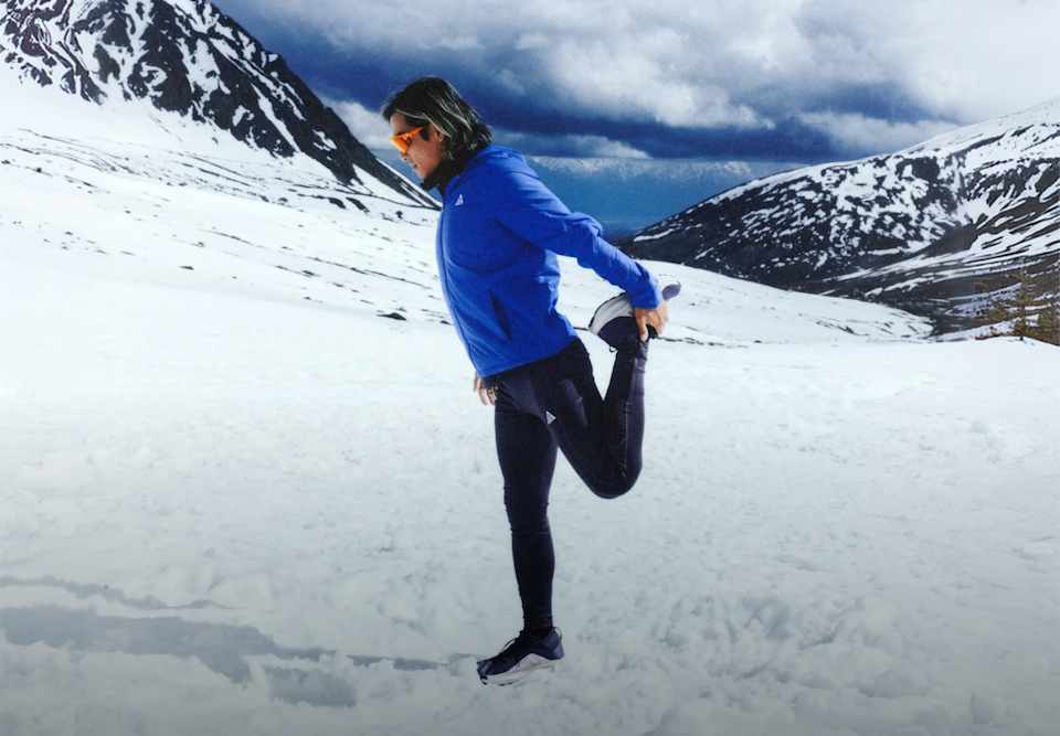 Person in blauer Nike ACG Jacke und schwarzer Leggings dehnt den Oberschenkel in einer weitläufigen verschneiten Berglandschaft., Wo der Asphalt endet, beginnt dein Lauf.