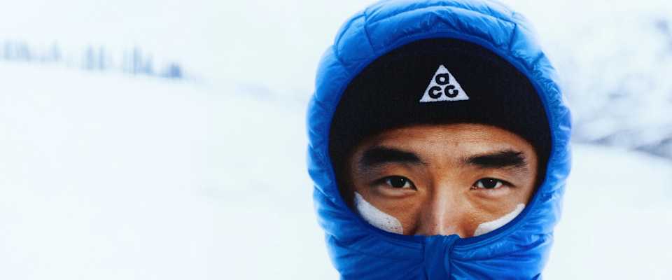 Mann blickt in verschneiter Umgebung nach vorn, mit weißer Creme auf den Wangen, blauer Nike ACG Lava Flow Steppjacke und schwarzer ACG Mütze.