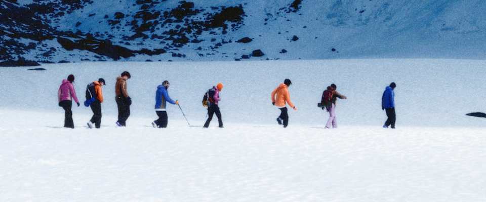 Un groupe de personnes portant des vestes Nike ACG Lava Flow colorées randonnent dans un champ de neige sous une imposante montagne enneigée.