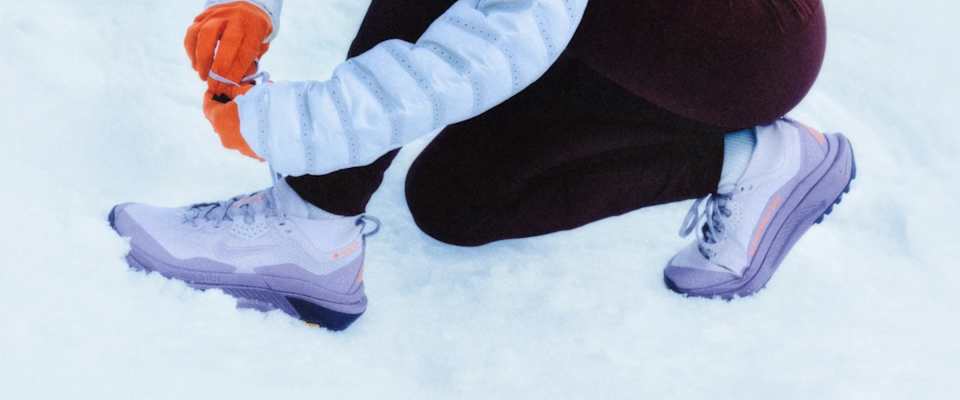 Gros plan sur une personne aux gants orange qui lace sa chaussure de trail Nike ACG violet clair dans la neige.