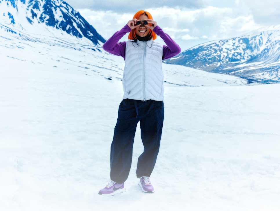 Lila, hosszú ujjú felső felett fehér Nike ACG mellényt viselő, mosolygó, élénk narancssárga hajú ember egy kis távcsővel a kezében, havas hegyi háttérrel. , Szabályozd a hőmérsékleted
