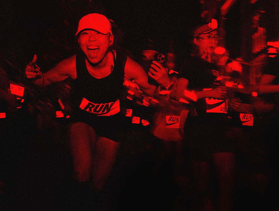 A woman smiling and running on a dark road at night, giving a thumbs-up, wearing a red cap and a black 'RUN' vest., Nike Experiences