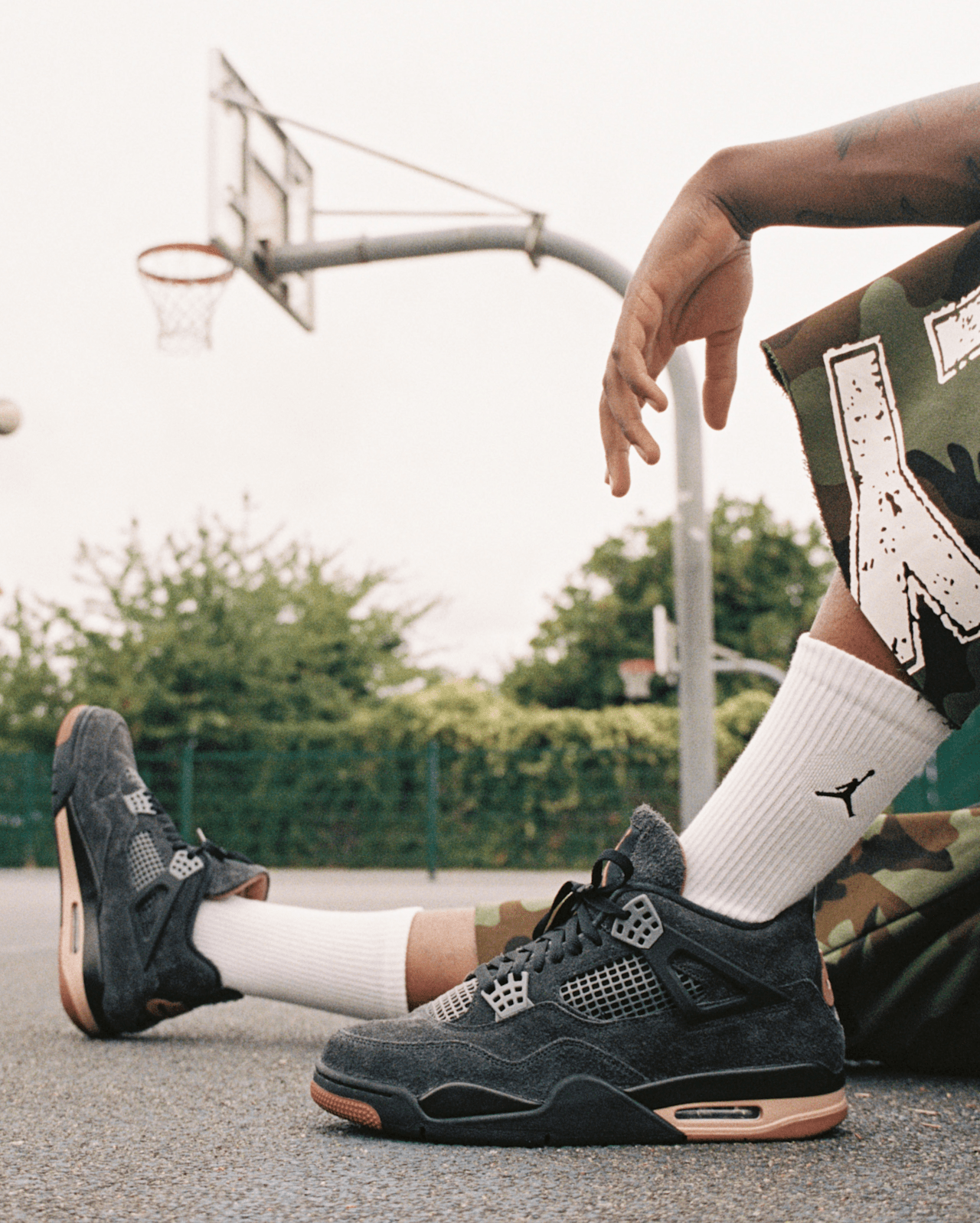 Un hombre con pantalón corto de camuflaje, calcetines blancos con logo Jordan y zapatillas Jordan 4 en gris oscuro con suela marrón.