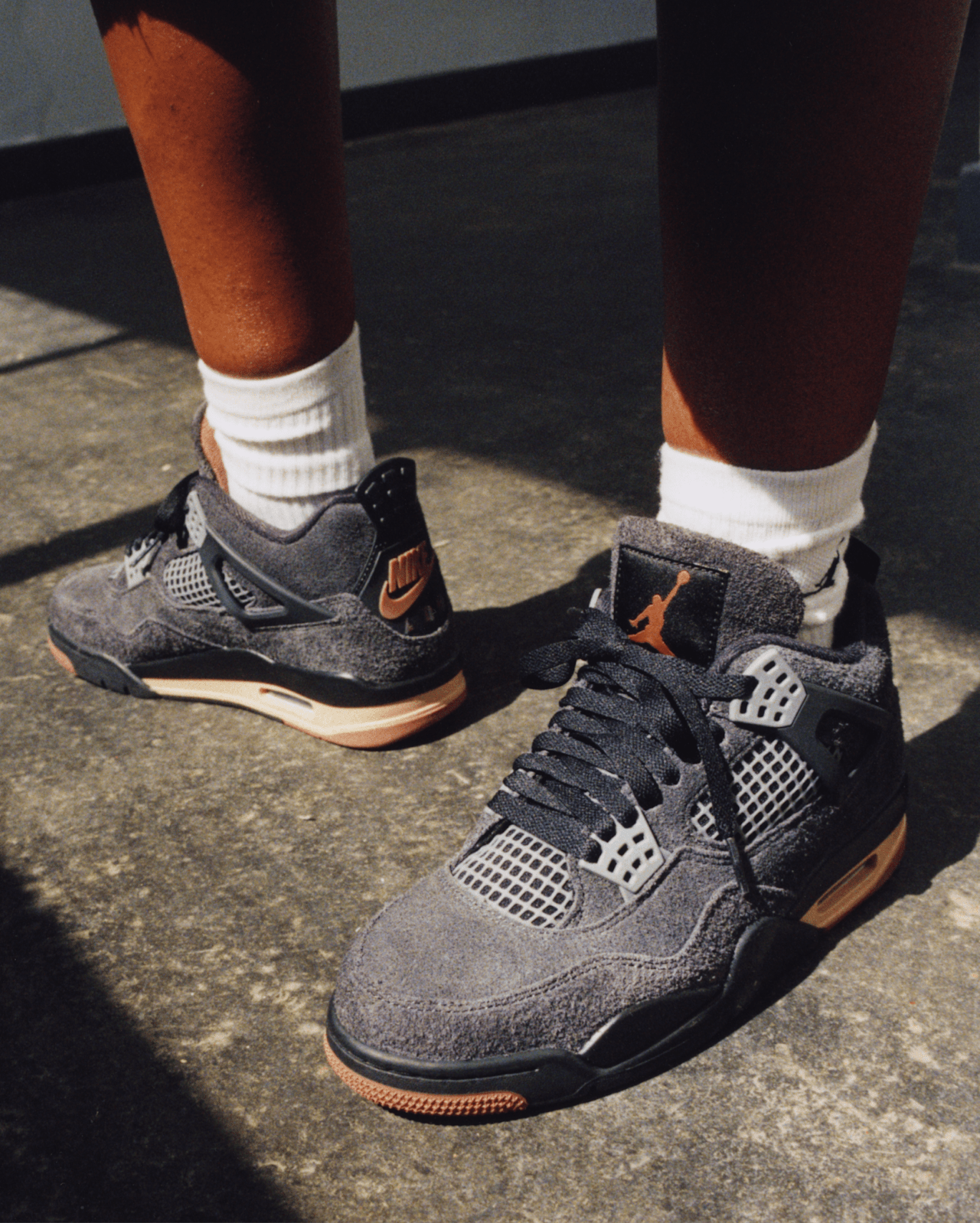 Iemand staat op een grijze betonnen ondergrond en draagt witte sokken en Air Jordan 4 sneakers in grijs, zwart en oranje.
